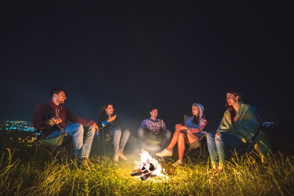 The Happy People Sit Near The Bonfire On The Background Of The City. Night Time