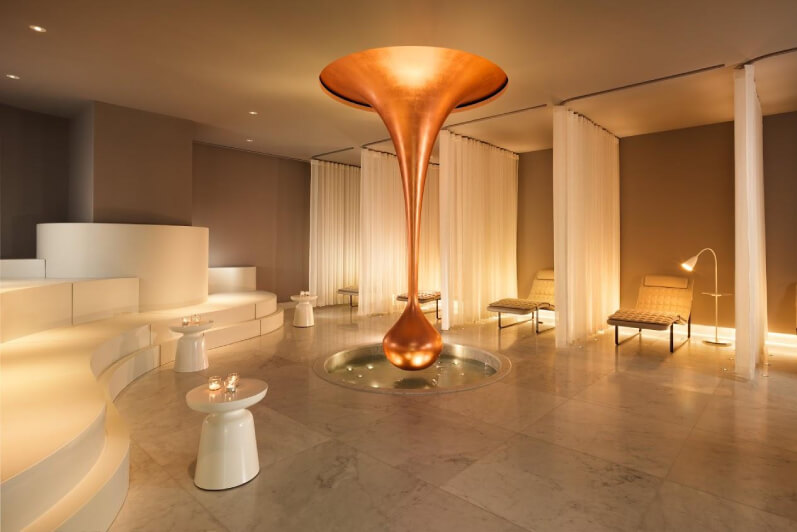 A modern white and golden spa with treatment chairs on the side, separated by curtains. There's a large piece of decor in the centre, looking like a golden drop dripping from the ceiling.
