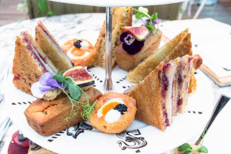 A platter of sandwiches with jam, boiled eggs in batter, and pieces of cake decorated with fresh fruit and flower petals.