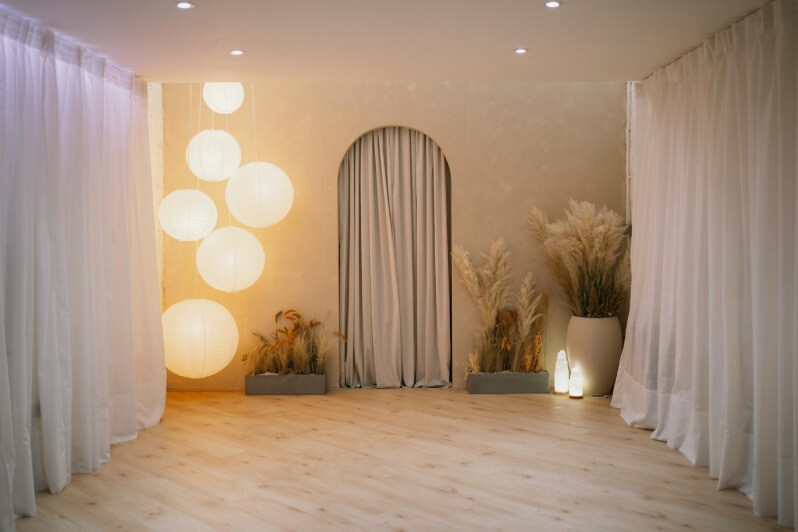 Calming studio with curtains on the side, an arch entrance in the centre, multiple warm light features, and plants on the floor.