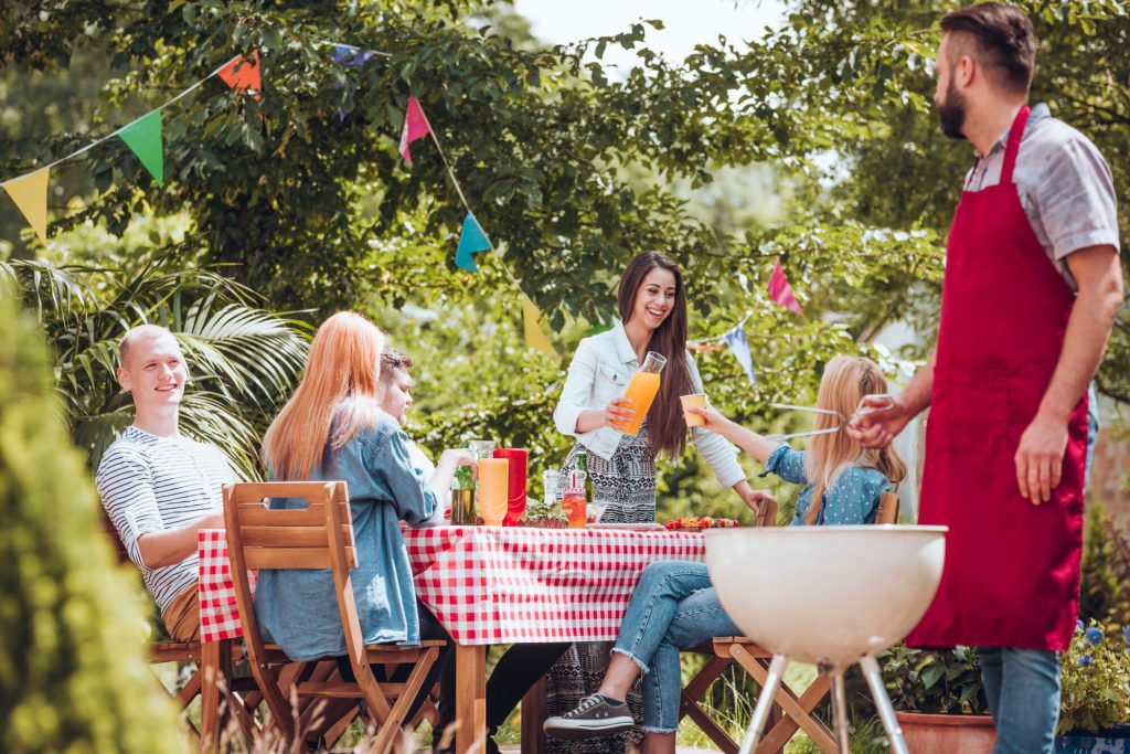 A group of people sitting at a table in a decorated backyard, enjoying some drinks and a BBQ.