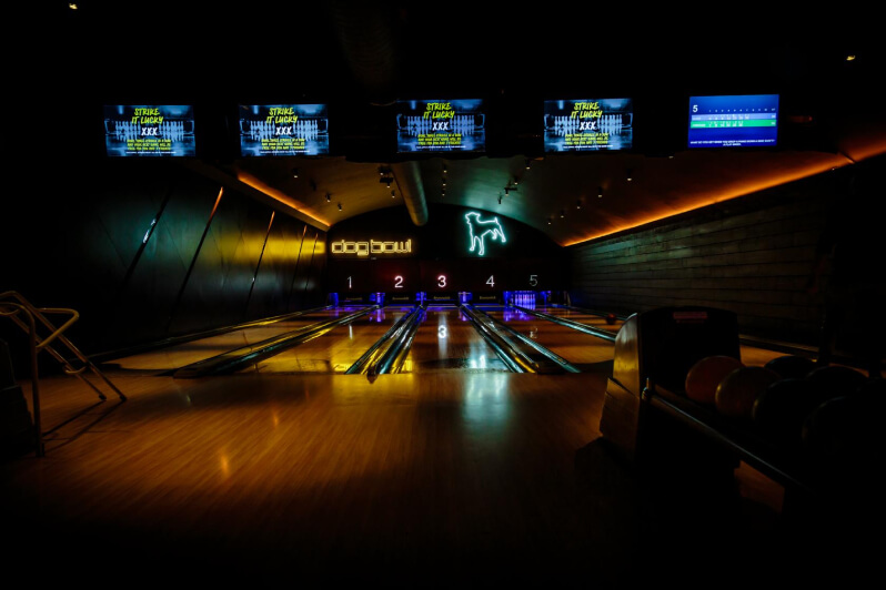 Dark bowling venue with ambient lights on the sides and neons over the alleys. There are five bowling alleys.