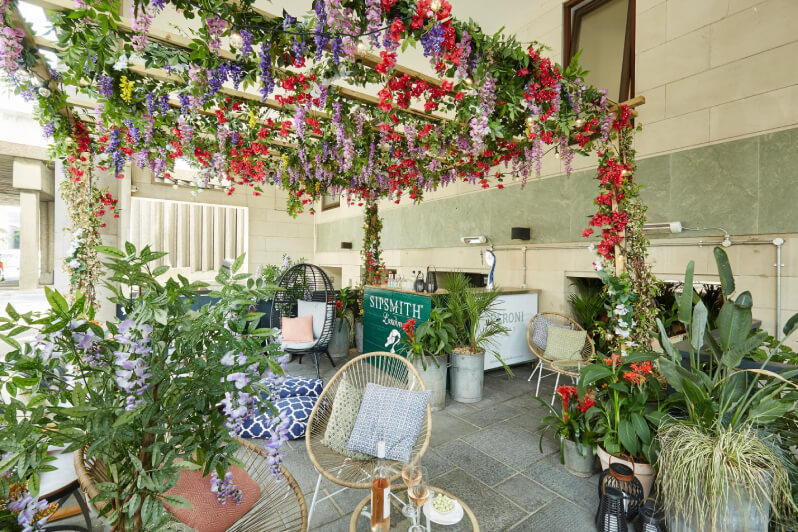 Shaded terrace with a small bar in the middle and oval garden chairs stood at cafe tables. THere are large plants stood in pots all over the place and flowers hanging from the ceiling.