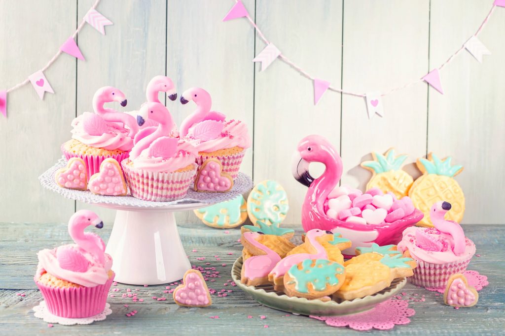 A table with flamingo-themed decorations, flamingo- and pineapple-shaped cookies, and cupcakes with sugar flamingos on top.