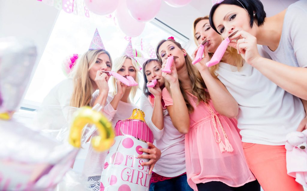 A group of women wearing party hats, blowing party horns, and posing with a baby-bottle-shaped balloon that says 