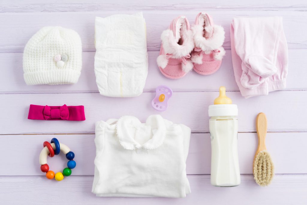 A flat-lay display with baby accessories: a hat, diaper, headband, rattle, pacifier, bottle, hair brush, clothes, and shoes.