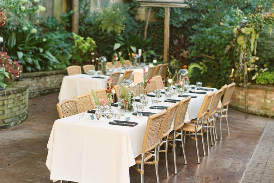 A long table covered with a white cloth set out in a private garden, surrounded by lush greenery.