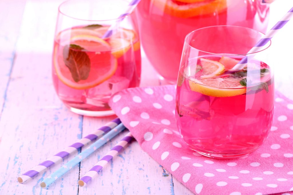 A pitcher and two glasses with pink-colored liquid, slices of lemon, mint leaves, and straws.