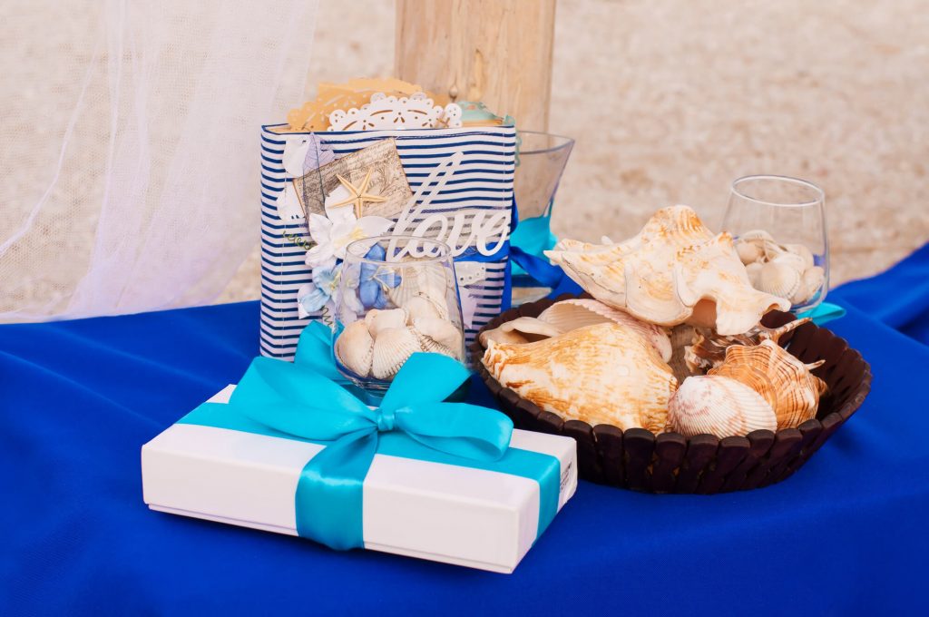 A table covered with a blue tablecloth, featuring a vase and glasses filled with seashells, a gift bag, and a wrapped present.