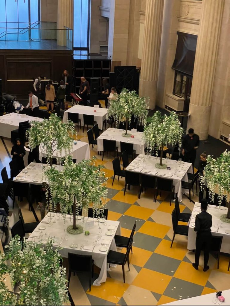 A wedding reception layout in Banking Hall in London with rectangular tables decorated with trees with white flowers and airy lights.