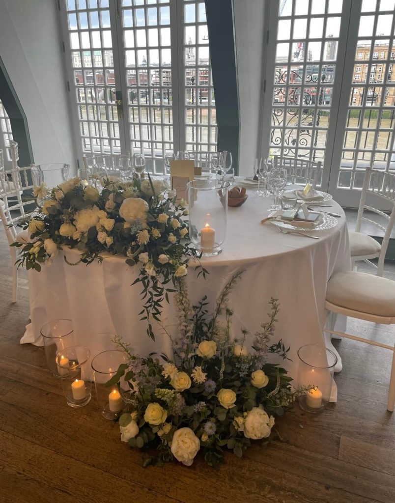 A round wedding top table with an open space opposite the couple's chairs, set with a white table cloth and decorated with white flowers and candles.