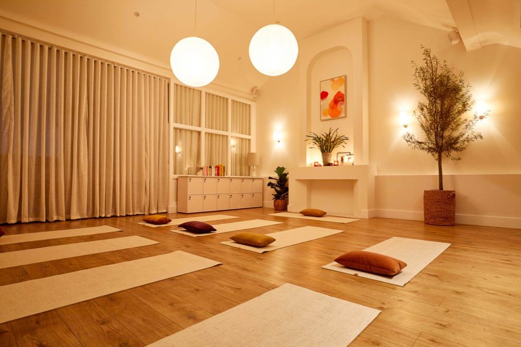 Serene yoga class setup in a warmly lit room with neutral-toned mats, plump cushions, and a calming décor featuring a potted tree and abstract wall art.