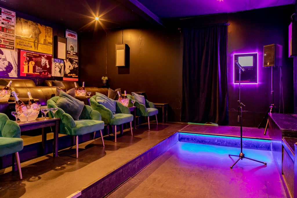 Intimate karaoke room with teal velvet chairs, a small stage with a microphone, and mood lighting, creating a perfect setting for a birthday singalong.