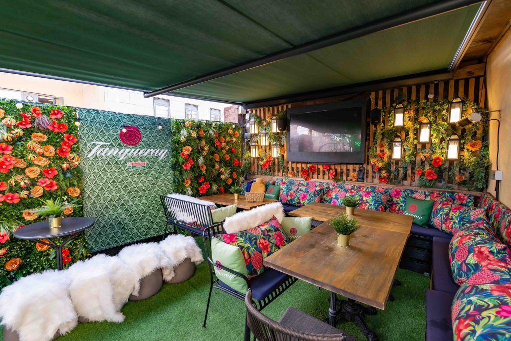 Vibrant rooftop terrace adorned with lush green walls, floral cushions, and a Tanqueray branded space, ideal for a festive outdoor birthday celebration.