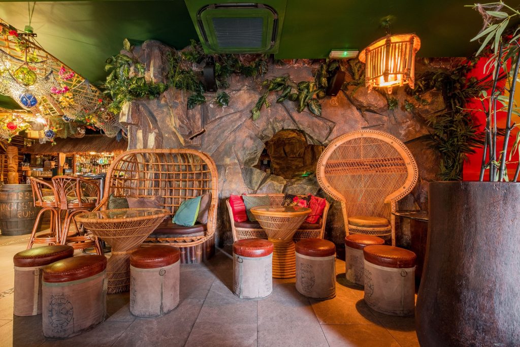 Eclectic jungle-themed section of a bar with wicker furniture, a stone cave, and vibrant plants for a tropical escape on your birthday.