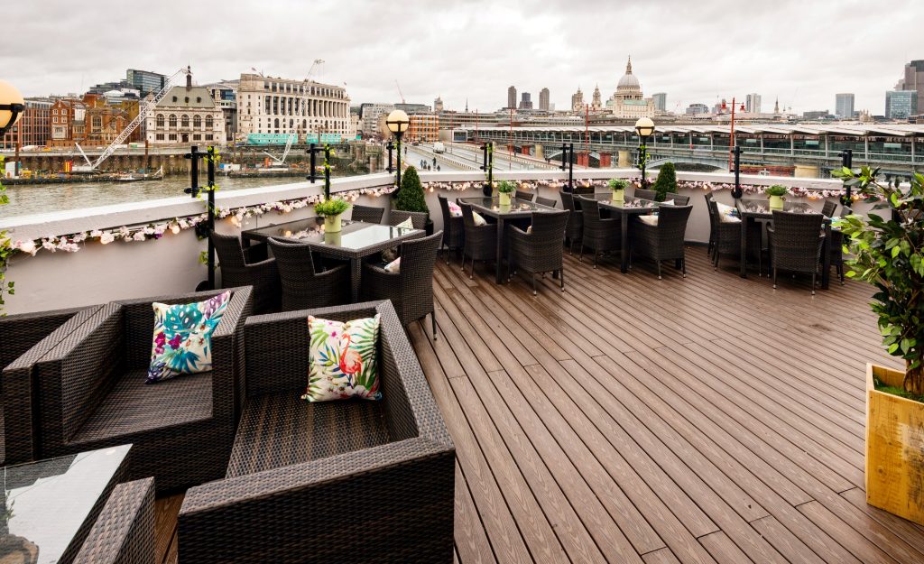 Riverside terrace with comfortable wicker seating, floral accents, and panoramic views of London’s architecture, perfect for a scenic birthday gathering.