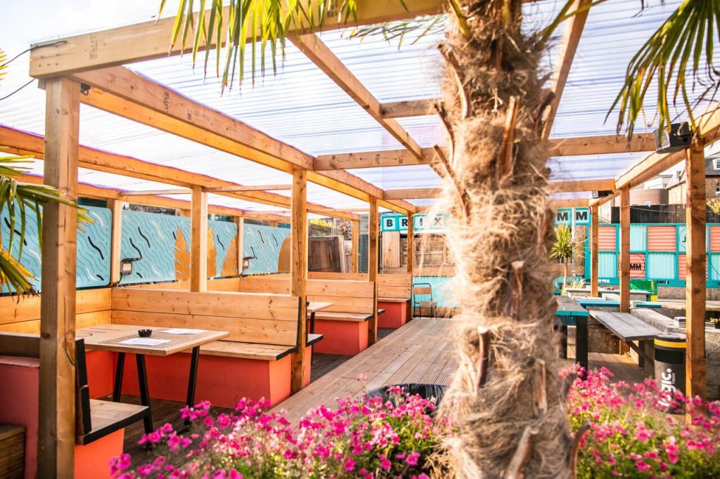 Sunny outdoor courtyard with wooden shelters, tropical plants, and colourful seating, providing a relaxed vibe for a summery birthday celebration.