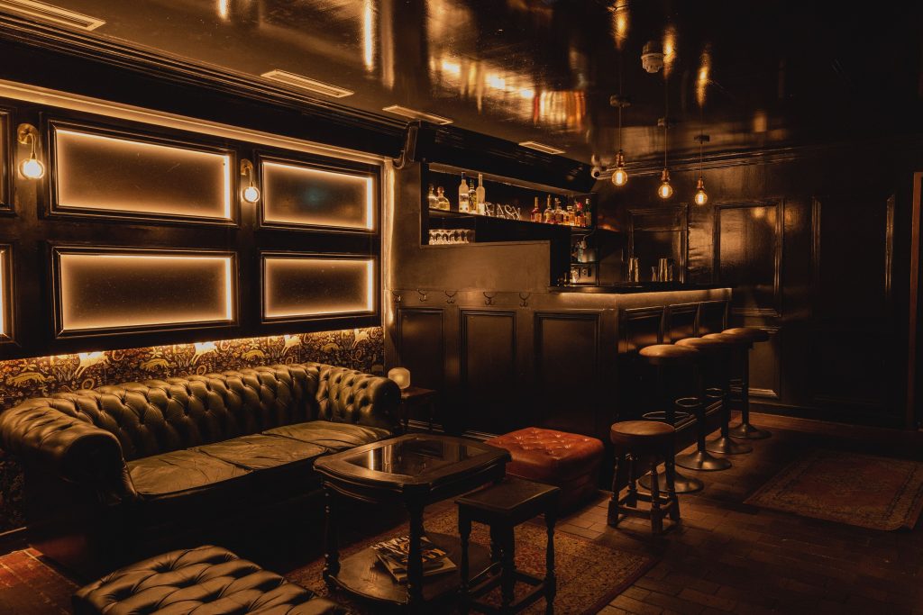 Moody vintage lounge with black tufted leather sofas, dim lighting, dark wood panelling, and a classic bar setup for a sophisticated, intimate gathering.