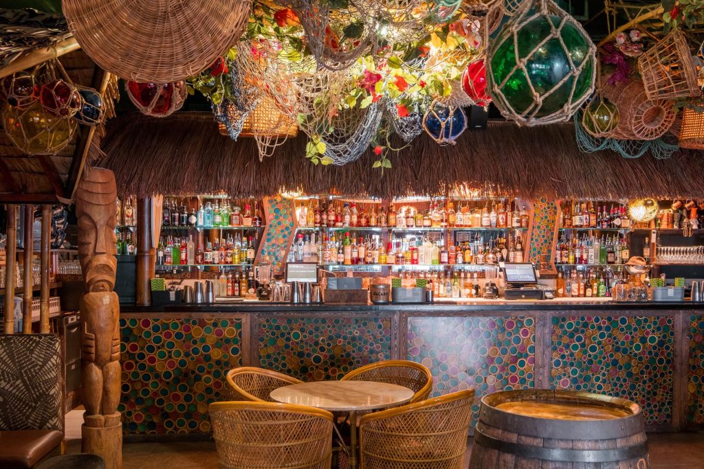 Cosy tiki-themed bar with an array of bottles on the shelves, fishing nets and colorful glass buoys hanging from the ceiling, and rattan furniture creating a tropical atmosphere.