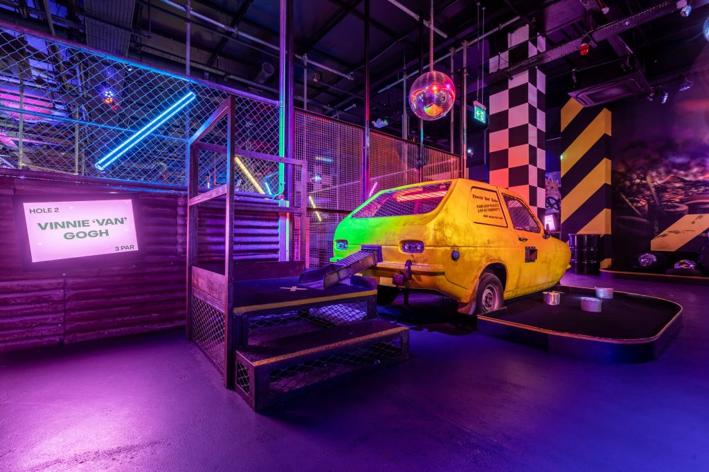 Quirky indoor mini-golf course with neon lights, a yellow car obstacle, and playful décor, offering a vibrant setting for a crazy golf birthday party.