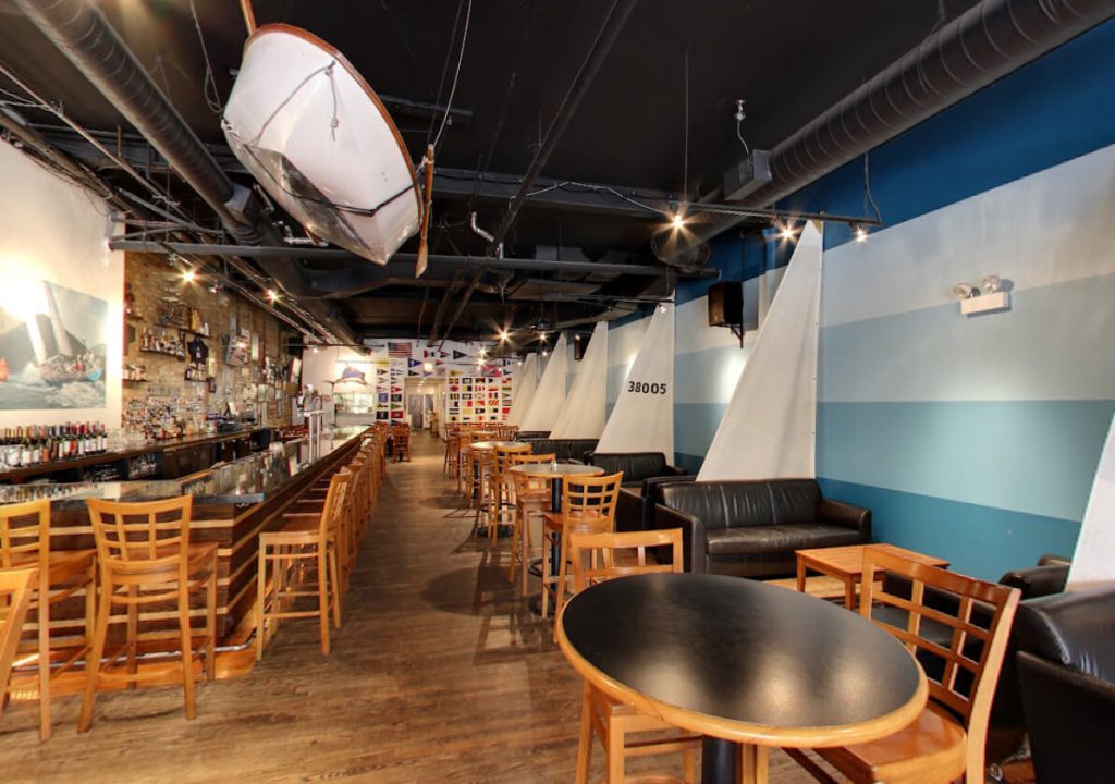 Nautical-themed Weather Mark Tavern in Chicago with a unique ceiling installation of a white sailboat, contrasting against a dark ceiling with exposed ductwork. The tavern has an extensive bar lined with wooden stools, sail-like dividers creating semi-private spaces, and maritime flags adding to the seafaring ambiance. The open layout with wood flooring and cozy seating offers a relaxed and adventurous dining experience.