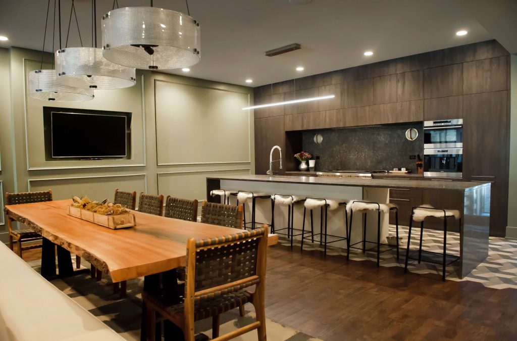 Modern and sleek dining space at BLOCK 25 in Chicago, featuring a long wooden communal table with stylish leather chairs, and elegant, circular crystal chandeliers. The kitchen sports a contemporary design with a marble countertop and state-of-the-art appliances, set against wood-paneled walls. The room's muted color palette and sophisticated decor create an atmosphere that's both upscale and welcoming.