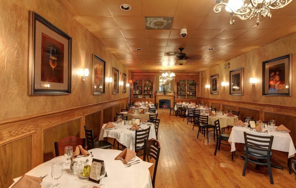 A cozy and elegant dining room in Bella Mia Fine Dining restaurant in Chicago, featuring neatly arranged tables with white tablecloths, brown napkins, and glassware. The room is adorned with framed artwork on warm, textured walls, a decorative chandelier, and a lit fireplace creating an inviting ambiance. The wooden floor reflects the soft lighting from wall-mounted fixtures, enhancing the room's classic charm.