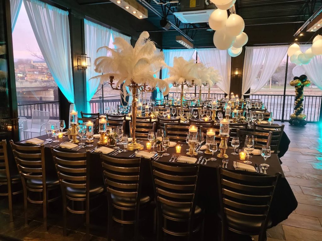 Elegant banquet setup in Eden Restaurant & Events in Chicago, featuring a long dining table adorned with white feather centerpieces, blue uplighting, and balloon decorations. The table is fully set with crystal glassware, silver cutlery, and lit candles, creating a festive and sophisticated atmosphere with a view of the city through draped windows in the background.