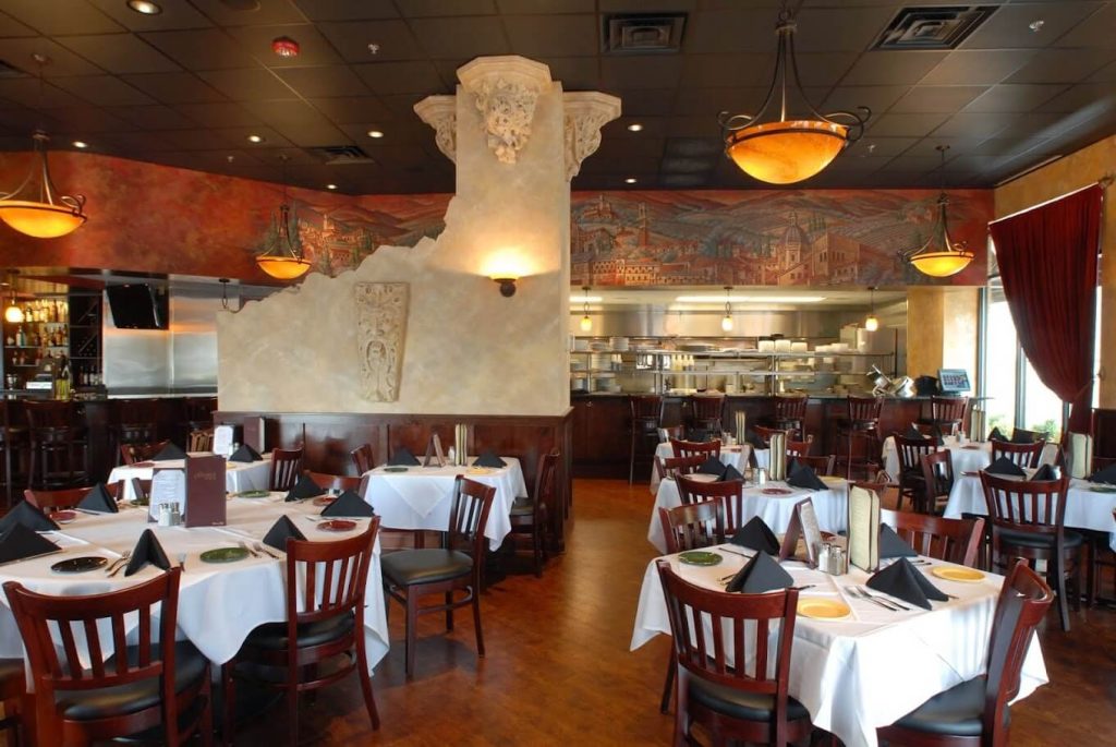 Traditional Italian dining room at Gianni's Cafe in Palatine, highlighted by a mural of an Italian landscape, ornate columns, and warm pendant lighting. The tables are draped with white tablecloths and set for service, reflecting the restaurant's classic charm. A view into the open kitchen adds an interactive and welcoming element to the dining experience.