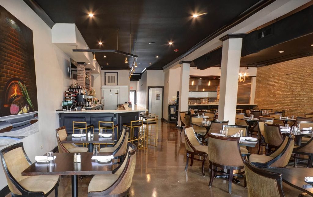 
Modern and stylish interior of La Parra Restaurant & Bar in Chicago, showcasing a spacious dining area with glossy concrete flooring, dark ceiling with recessed lighting, and a bar area with high stools. The walls are partially adorned with a large mural and exposed brick accents, complementing the contemporary dark wood tables and chairs with plush beige seating. Each table is set with white plates and silverware, ready for guests. The ambiance is upscale with a casual touch.