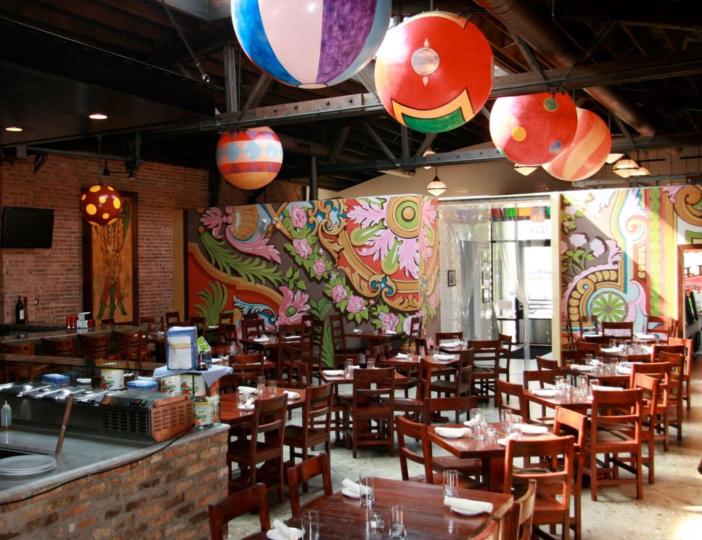 Vibrant and colorful dining area at Macello Cucina di Puglia in Chicago, featuring whimsical balloon-like light fixtures, exposed brick walls, and a lively mural with floral and abstract designs. Wooden tables and chairs are neatly arranged, ready for guests, contributing to the restaurant's lively and artistic atmosphere.