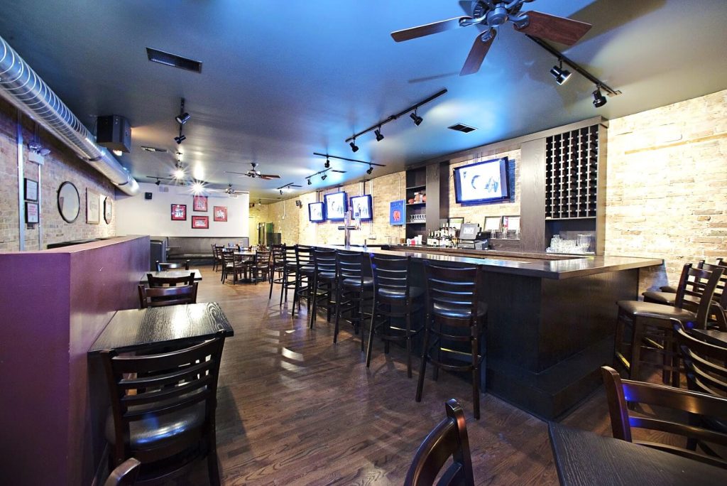 Casual and inviting interior of Pizzeria Serio in Chicago, featuring a spacious dining area with dark wooden tables and chairs, a long bar with black bar stools, and a brick wall accent. The area is illuminated by ceiling fans and spotlights, and multiple televisions are mounted for viewing, creating a relaxed atmosphere suitable for casual dining and socializing.