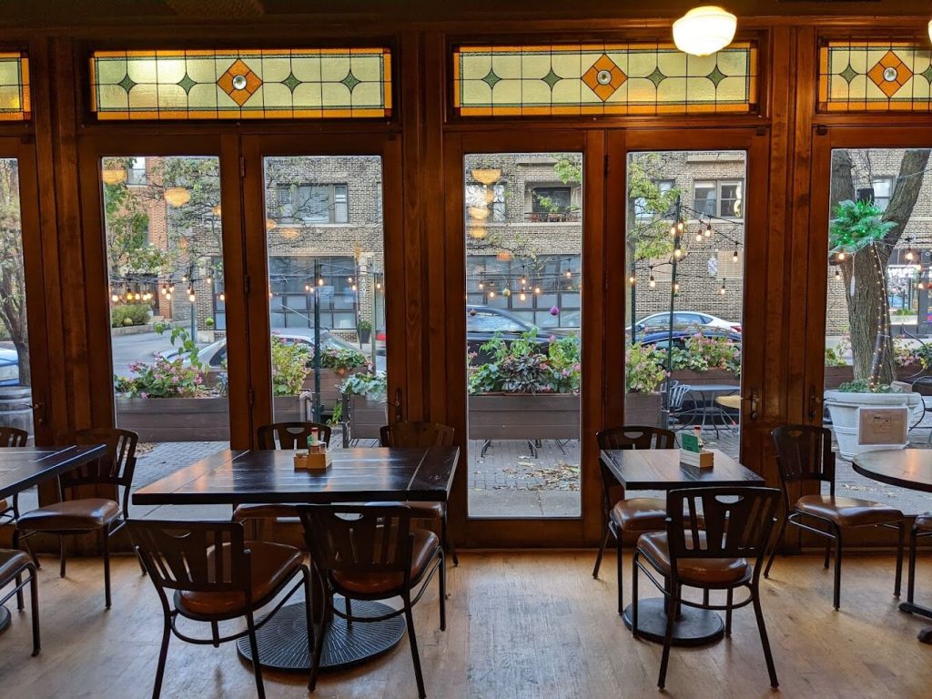 Cozy dining space at Port and Park Bistro in Chicago, with natural light streaming through large windows with stained glass accents. The room is furnished with dark wood tables and chairs, and pendant lights hanging from a high ceiling, offering a view of a busy street. The simple, yet inviting decor creates a pleasant atmosphere for casual dining.