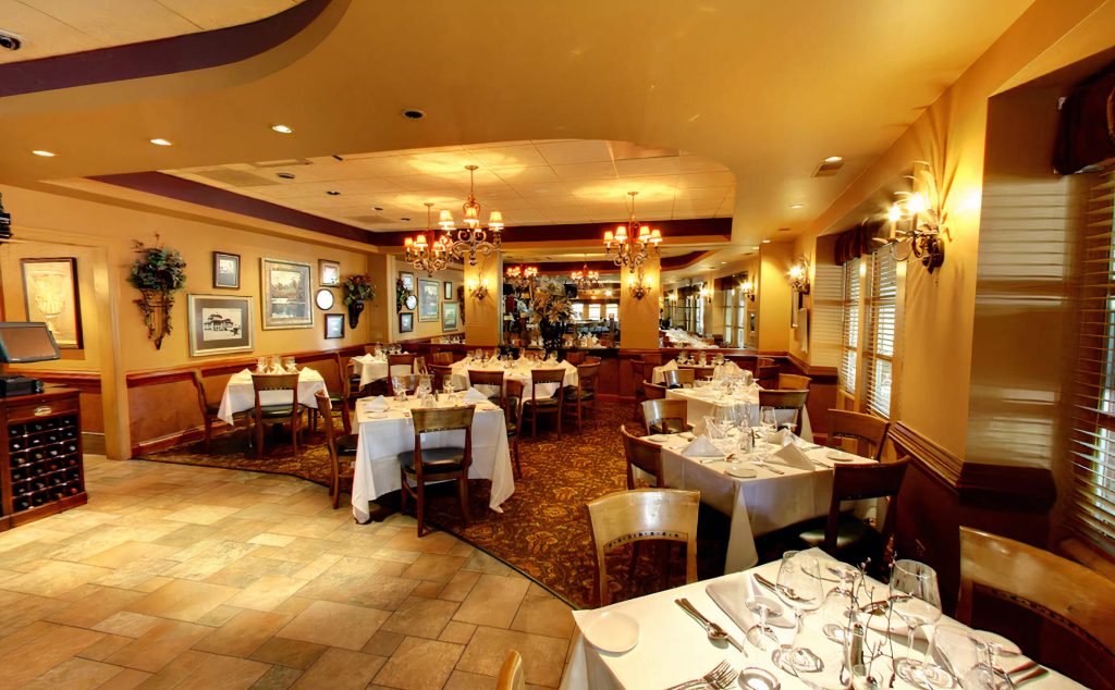 Classic and warm dining space at Roberto's Ristorante & Pizzeria in Chicago, with multiple tables set with white linens, wine glasses, and folded napkins. The room is lit by ornate chandeliers and wall sconces, with decor that includes framed pictures and wine storage, all complemented by a patterned carpet and stone tile flooring for a traditional Italian dining experience.
