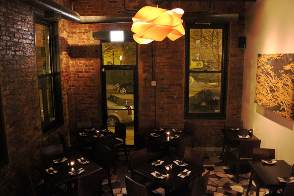 Intimate and ambient sushi bar at Seadog Sushi Bar in Chicago, featuring exposed brick walls and warm wooden floors. The dining area is lit with a soft glow from a unique, sculptural overhead light, and the tables are neatly set with black napkins, creating a cozy and modern dining experience.