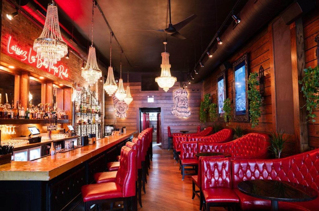 Chic and vibrant bar at Storyville in Chicago, with lustrous red tufted booths and bar stools, dark wood flooring, and an eclectic mix of ornate chandeliers. The walls are adorned with whimsical art and neon signs, while lush green plants add a touch of freshness to the lively and opulent New Orleans-inspired setting.