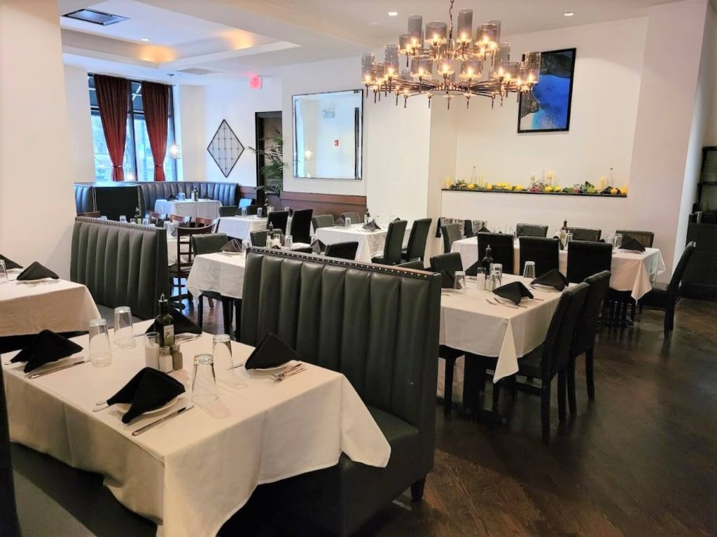 Elegant and modern dining room at Thassos Greek Restaurant in Clarendon Hills, featuring a bright space with large windows draped in heavy curtains, grey upholstered booths, and white tablecloth-covered tables. The room is lit by a stylish chandelier that adds a sophisticated touch to the clean and minimalist decor, inviting a comfortable yet upscale dining experience.