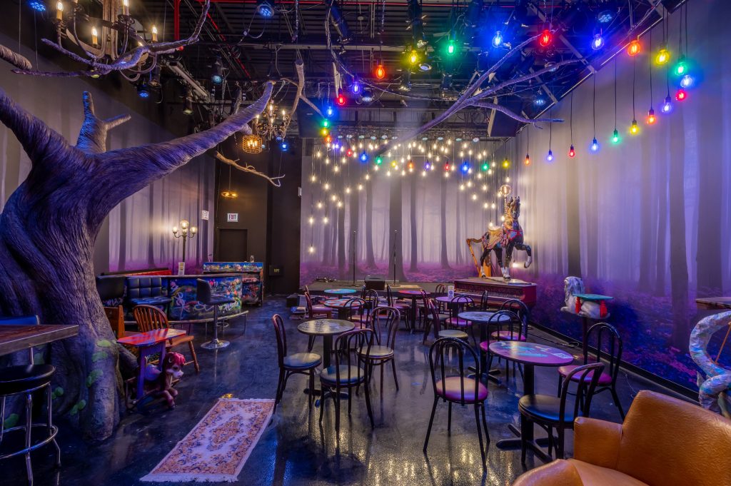 An eclectic and fantastical dining space at Whim at Stage 773 in Chicago, featuring whimsical decor with a large, artistic tree sculpture and a colorful carousel horse. The room is adorned with a canopy of bright, multicolored stage lights and string lights, creating a magical and theatrical ambiance. The varied seating, including cozy sofas and traditional chairs around intimate tables, is set upon a shiny floor that reflects the vibrant lighting, all contributing to a playful and immersive dining experience.