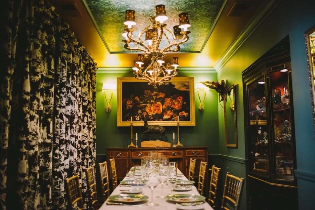 The Savarin Room at Marchesa in Chicago offers an opulent dining experience with its rich green walls, ornate golden chandelier, and classic paintings. The long dining table set with fine china and crystal glasses under the warm glow of candlelight creates an intimate and luxurious atmosphere reminiscent of a bygone era.