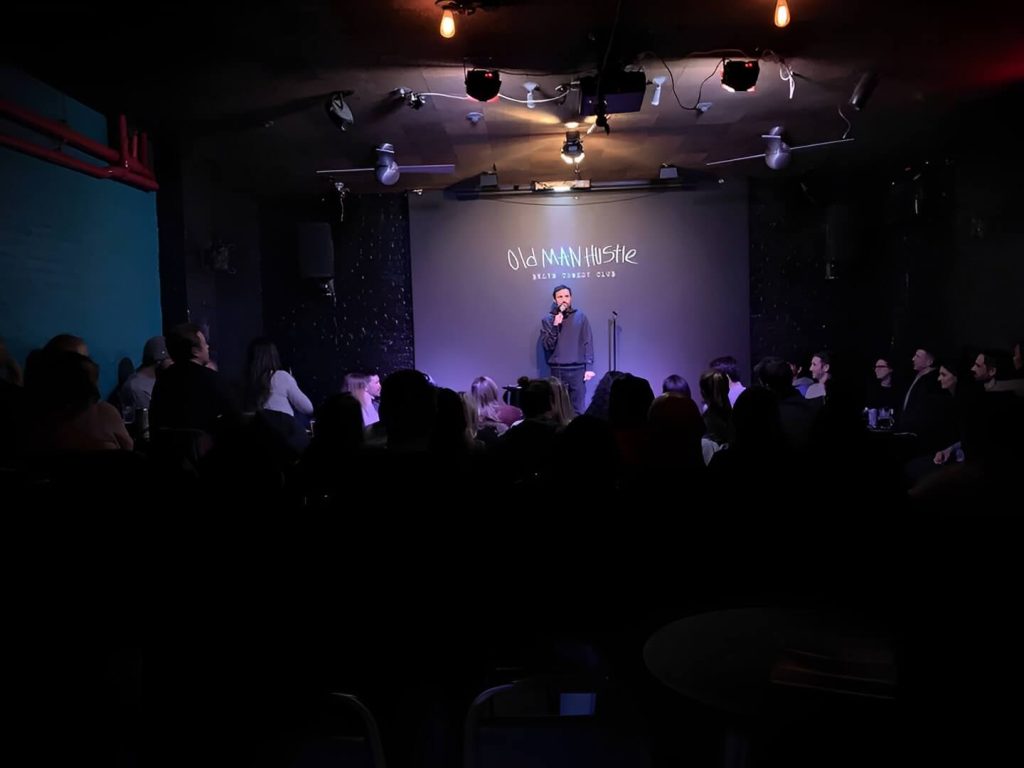 A comedy club with a performer on stage and an audience seated in a dark room, focused on the entertainer, showcasing a live performance environment.