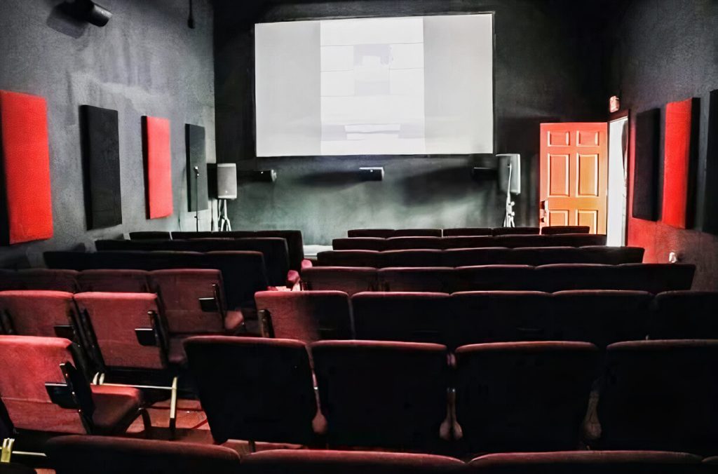 An empty movie theater with rows of red seats facing a blank screen. Acoustic panels, some red and some black, line the dark walls on either side, along with two speakers placed symmetrically. There's a visible exit door on the right-hand side of the room, painted in contrasting orange and white, providing a pop of color against the darker interior. The scene conveys a quiet ambiance, possibly before or after a film screening.