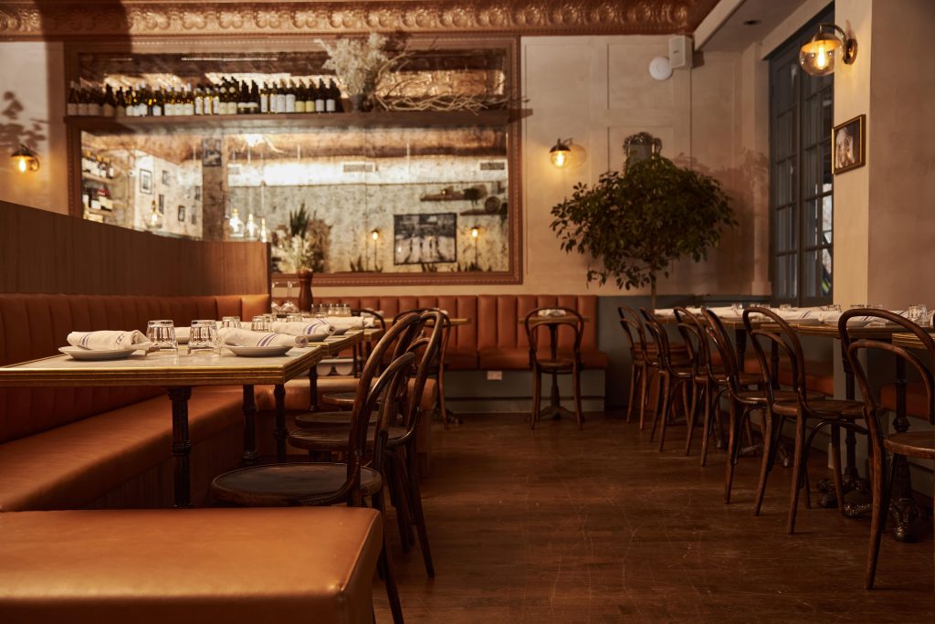 A restaurant with a cozy ambiance featuring a long dining table with a brown leather booth, wooden chairs, and elegant table settings, complemented by soft lighting and mirrored wall decor.
