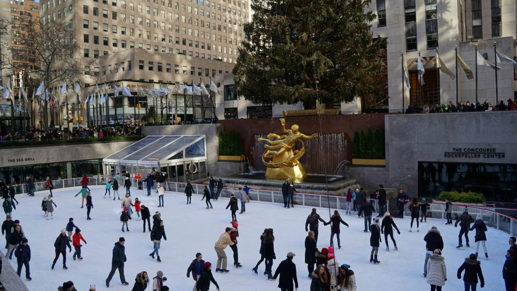 A bustling outdoor ice skating rink at Rockefeller Center during the holiday season, with skaters of all ages, a large Christmas tree, and the iconic golden Prometheus statue, offering a festive winter activity.
