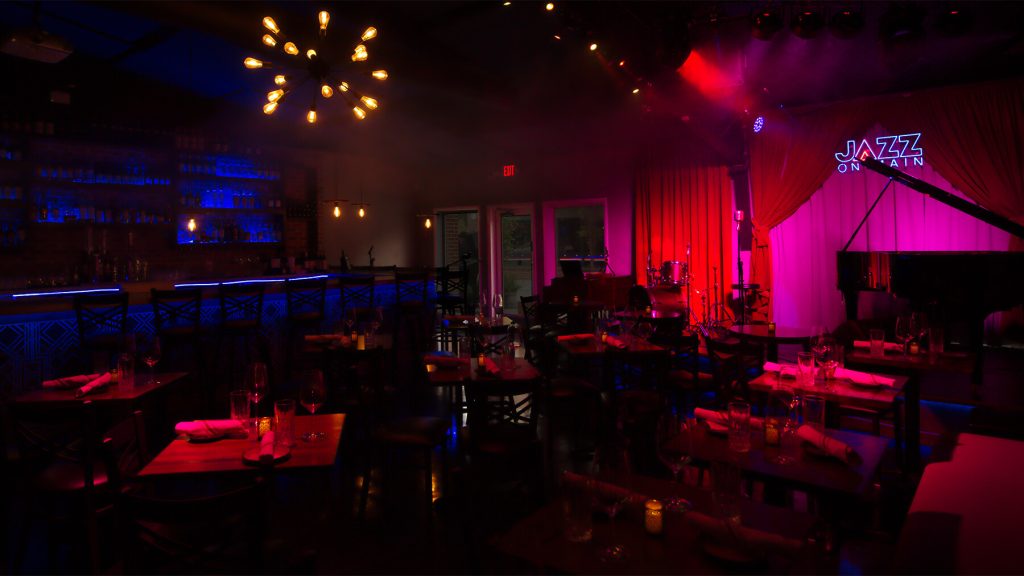 A jazz club with blue and red lighting, modern decor, and a stage set for a live performance, offering an intimate musical dining experience.