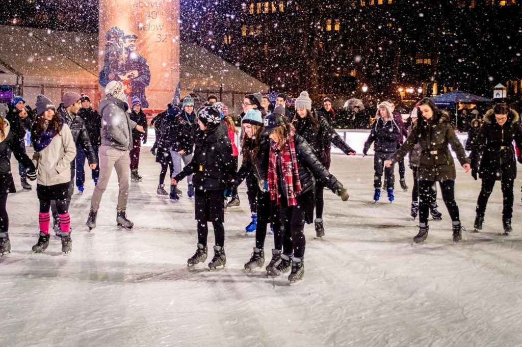 People ice skating on an outdoor rink at night during snowfall, with a large statue in the background and city buildings illuminated, creating a festive winter scene.