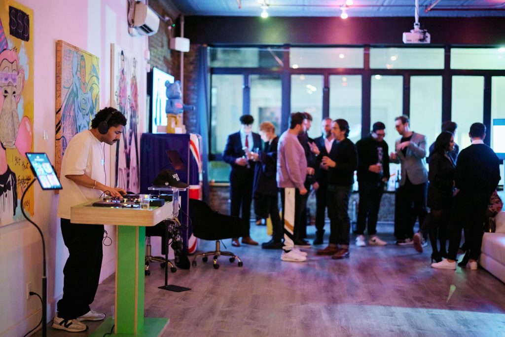A DJ is operating a turntable at an art gallery event, with colorful artwork displayed on the walls behind him. A group of guests is socializing in the background near large windows.