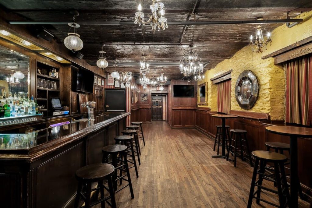 A rustic speakeasy-style bar with dark wooden paneling, classic bar stools, and vintage chandeliers, creating an old-fashioned and cozy atmosphere.