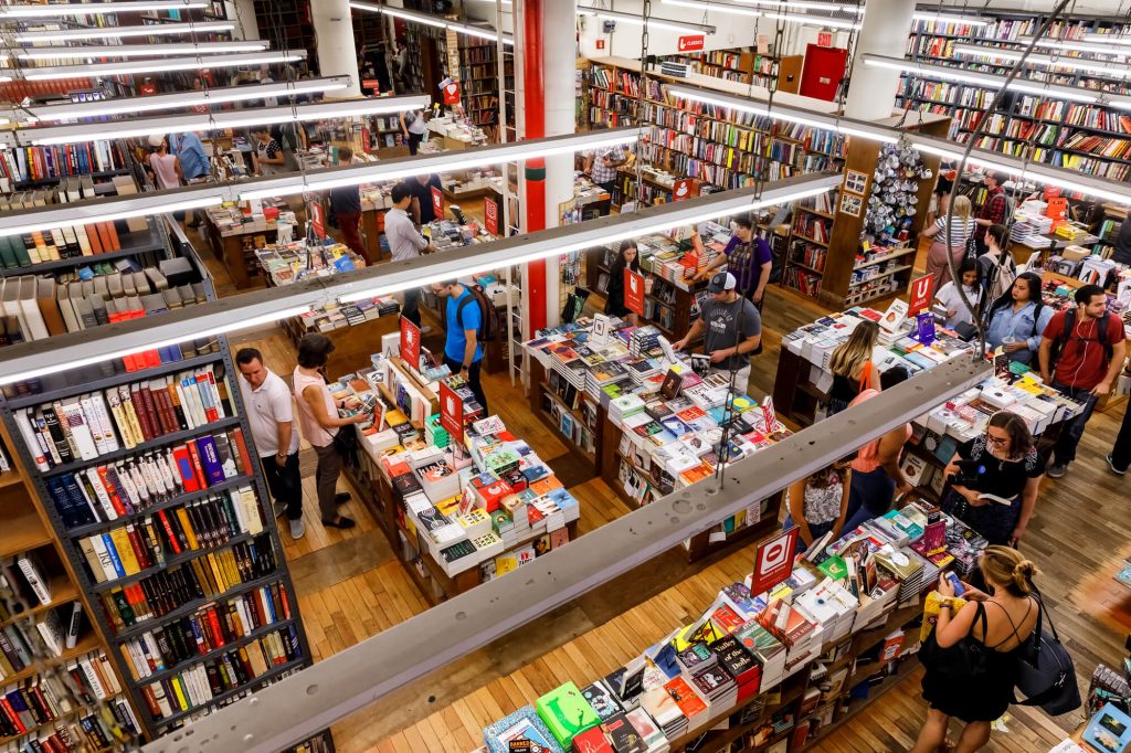 A bustling bookstore with multiple rows of bookshelves and customers browsing through an extensive collection of books. The view is from an upper level looking down on the main floor with a central display of books.