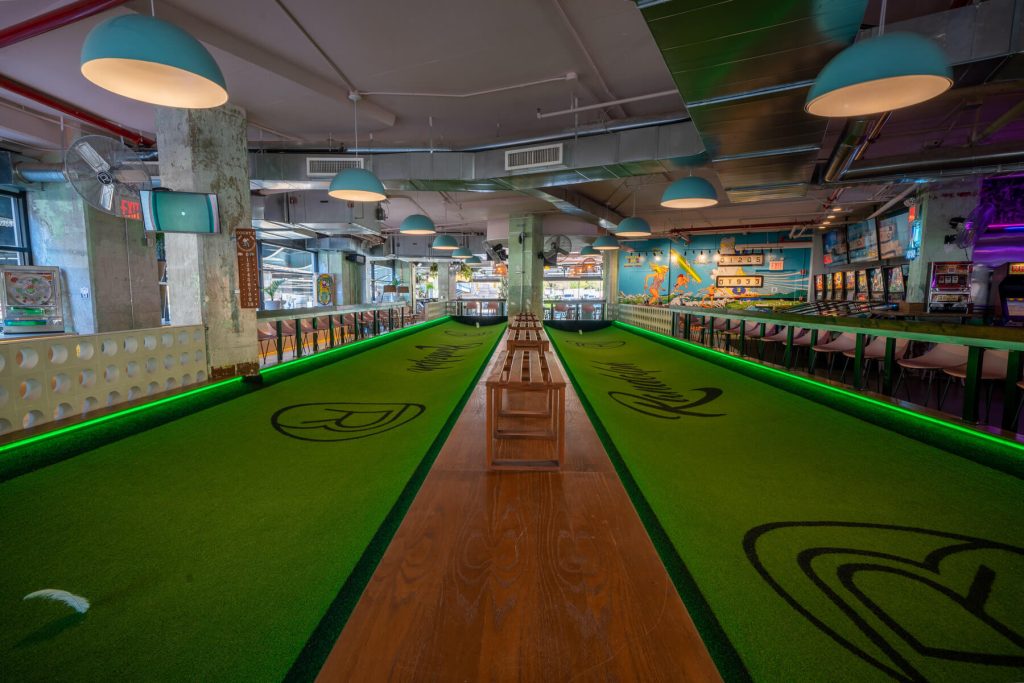 An indoor bowling alley with two lanes, one showing a close view of the wooden floor leading up to the pins, with neon-lit walls and hanging dome lights, creating a lively atmosphere.