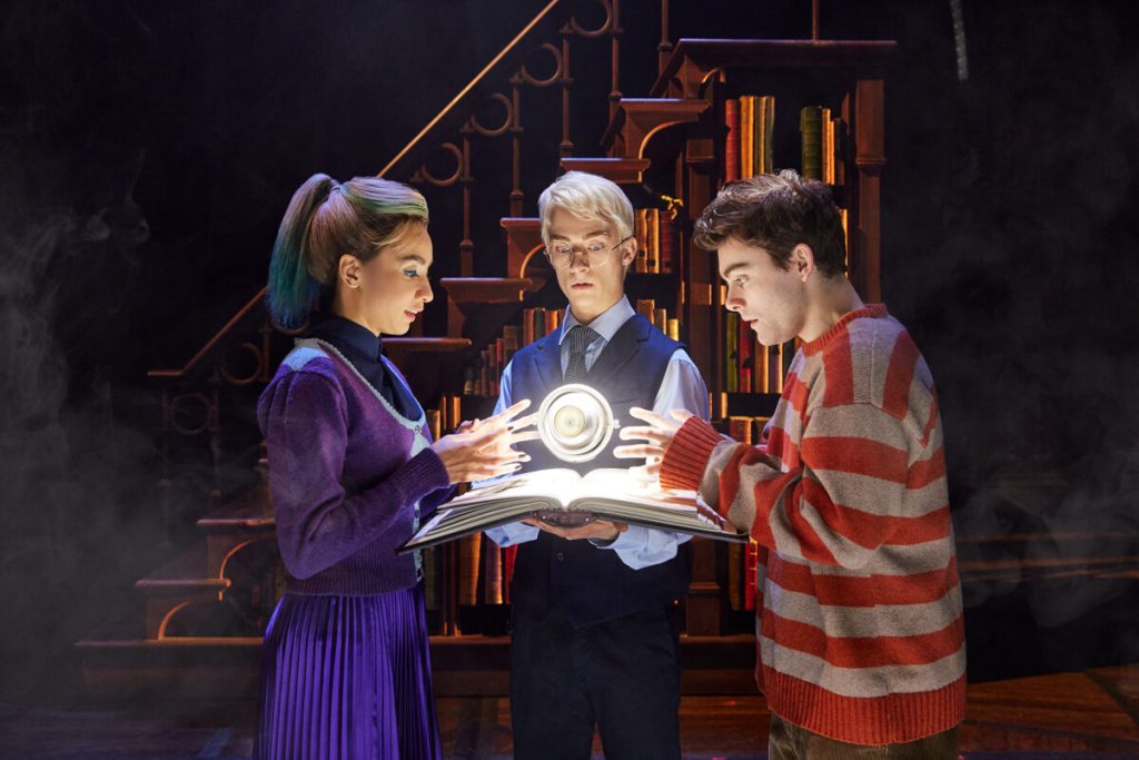 A theatrical performance with three actors, two of whom are looking in awe at a glowing book held by the third, set against a backdrop of a magical library, indicative of a family-friendly Broadway show.